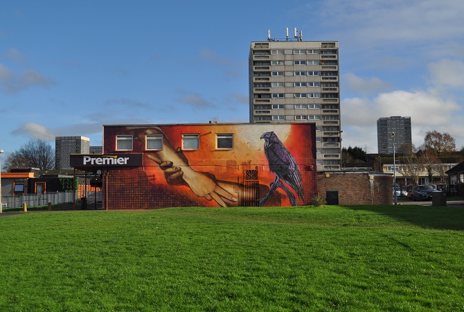 A Mural for Druids Heath at the end of a row of shops. A block of flats is in the background.