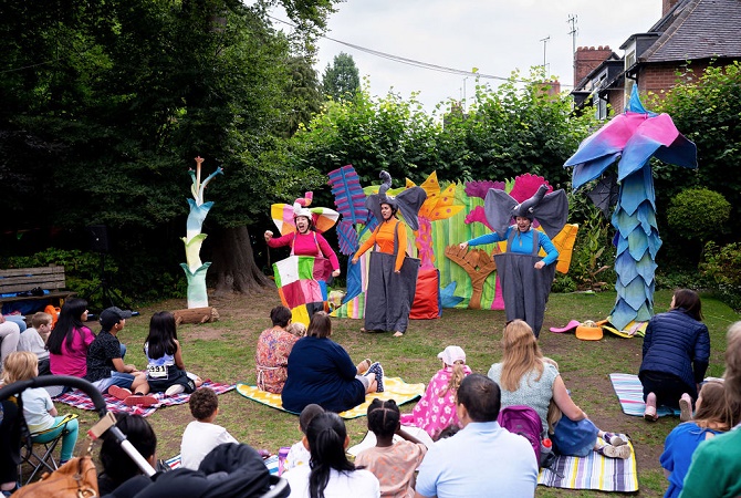 A group of actors performing Elmer’s Walk in colourful costumes in front of a family audience outdoors