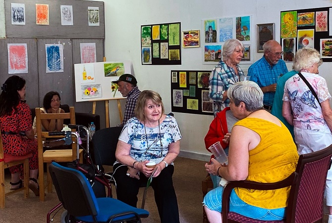 Two women sat down on chairs at the Erdington Creative Arts Programme. Behind the two women a group of people look at pictures of various pieces of artwork