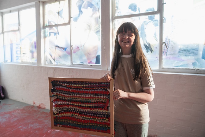 A woman holds up a tapestry creation in her hands